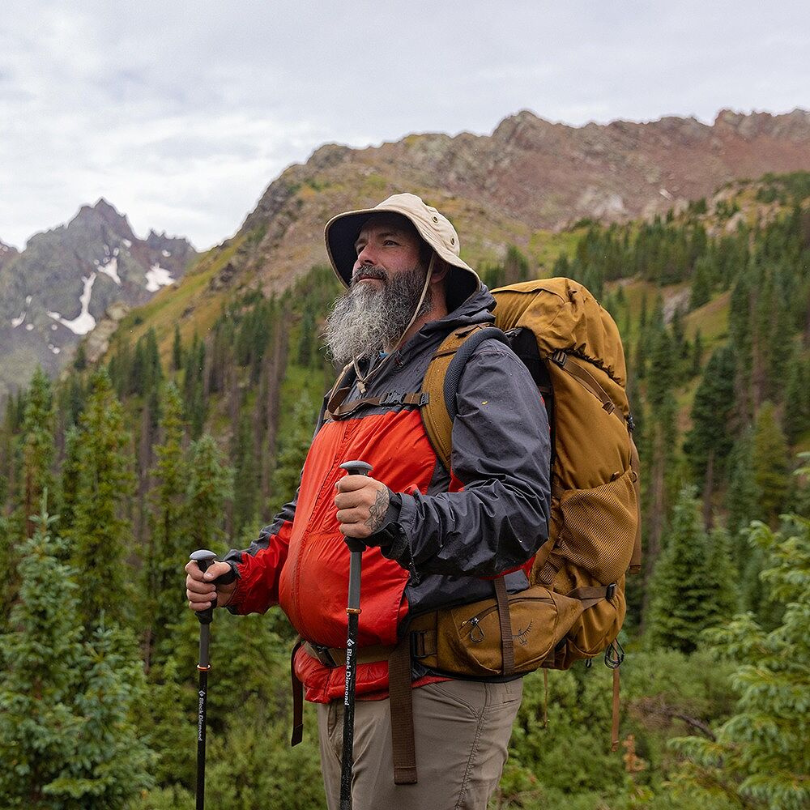Man with a backpack and hiking poles standing in a mountainous forested area.