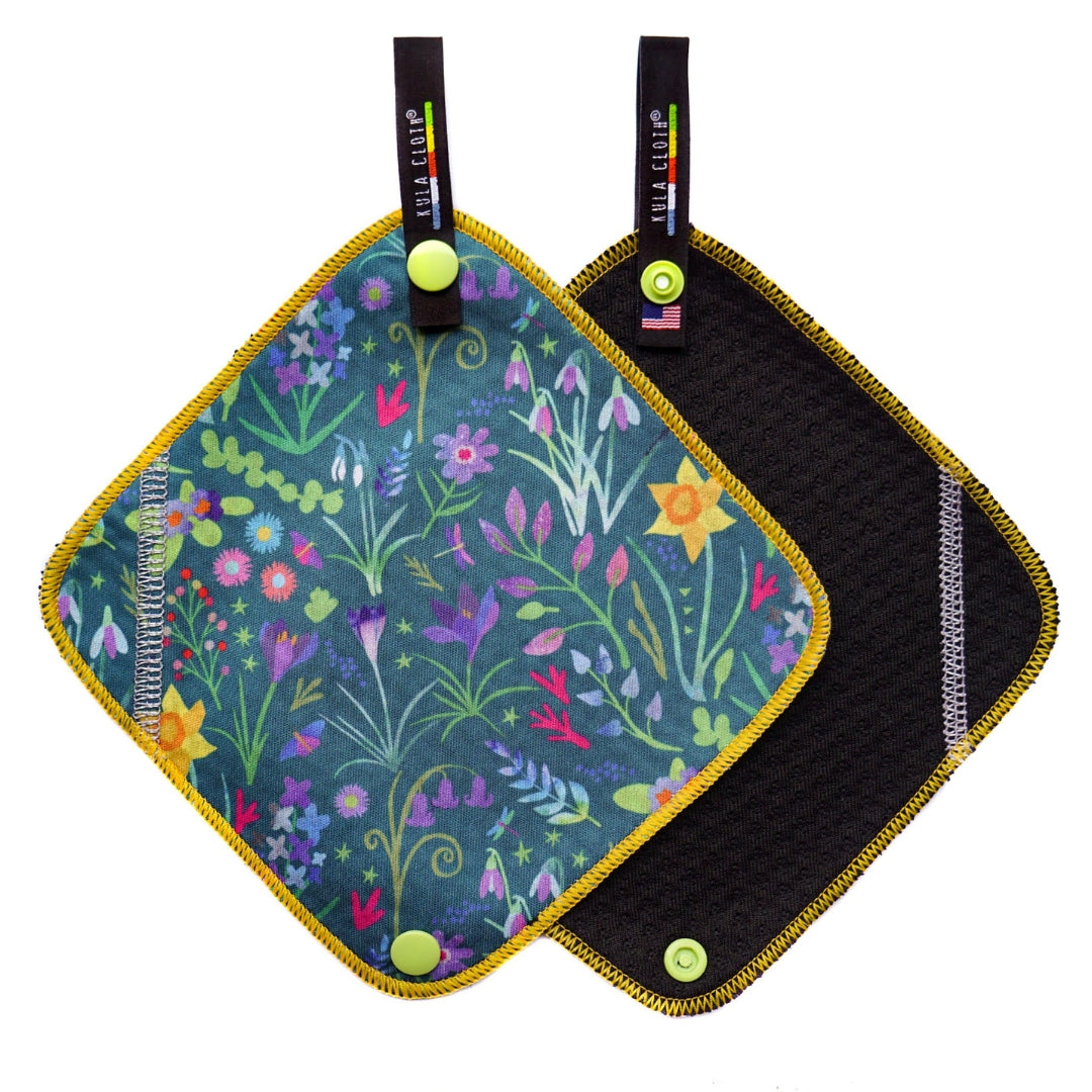 Two Kula Cloth pee cloths displayed against a white background, one with a floral pattern and the other in a solid black color, both with yellow trim and green snap buttons.